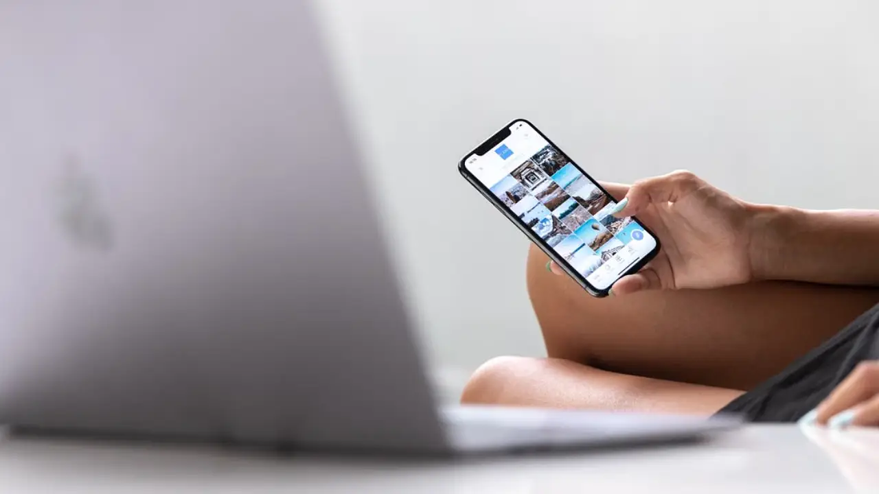 A person holds a smartphone showing a grid of posts beside an open laptop